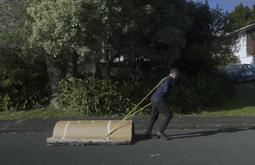 A person drags a roll of cardboard along a road with a yellow strap over their shoulder.