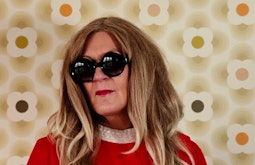 A person stands in front of a 60s wallpaper wearing a red dress, large black glasses, and a long blonde wig.