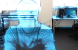 A person appears like a hologram in an office, they're made of translucent blue light.