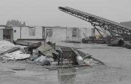 Detritus and broken objects sit in a concrete loading area. Run down buildings in the background. A large conveyer belt sits in disrepair.