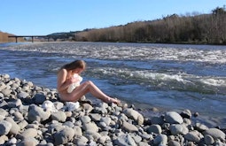A person sits naked on river-side rocks while holding a crumpled white sheet.