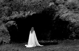 A person wearing a white sheet walks towards a large shadowy cave.