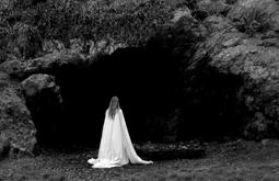 A person wearing a white sheet walks towards a large shadowy cave.