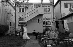 A person wearing a white sheet looks at a dog while standing in a backyard. There are chairs and a couch on the lawn.