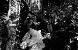 A person sits on the ground surrounded by bush while holding a white sheet.