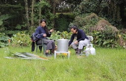 Two people sit in plastic chairs on a lush patch of grass. Dense bush surrounds them. There is a large metal pot and gas canister between them.