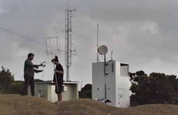 Two people stand by a communication tower holding radio equipment and headphones.
