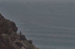 A person stands at a cliff-top look out facing towards the open sea.
