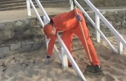 A person in an orange jumpsuit picks cleans a sandy beach, they are holding a rake.