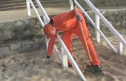 A person in an orange jumpsuit picks cleans a sandy beach, they are holding a rake.