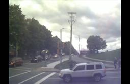 At an intersection cars wait, people walk by. A large row of trees runs beside the road.