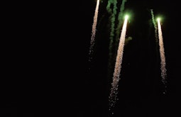 Three fireworks with spark trails shoot upwards on a dark night.