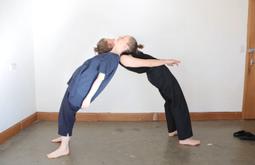 In a white walled room two people lean backwards against each other, their head resting on the others shoulders.