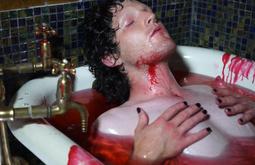 A person lies in a bath holding their chest, the water is red with fake blood. There is blood running down their neck and the side of the bath.