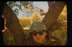 A man leans against a tree wearing a army green helmet and sunglasses.