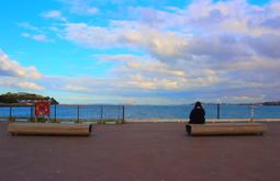 A person sits on a bench by the seaside. There is a matching empty bench nearby. There is bright blue sky with some clouds above.