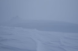 A landscape of snow, there is a hill in the distance through the fog.