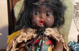 A doll with large black eyes and red lips wears customary Māori attire, there is a tiki hanging from the dolls neck.