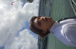 A person looks at the camera with their mouth slightly agape, they're wearing a white shirt and tie. There is a large cruise ship in behind them.