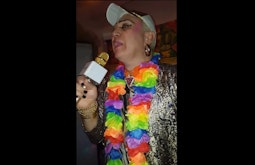 A person wearing a rainbow lei and gold hat sings into a golden microphone.