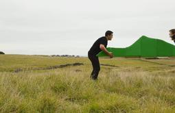 In a large field two people carry a green sculpture of a hill.