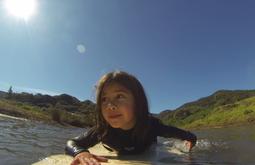 A child is lying down on a surfboard while swimming out in water. The sun and blue sky are above them.