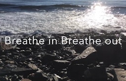 In low sunlight a small waves breaks on rocks. Over the top is the text 'Breathe in Breathe out'.