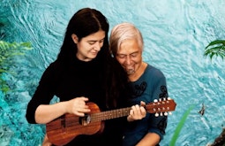 A person sits on their parents knee while playing the ukelele. A bright blue body of water is super-imposed as the background.