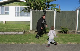 A person runs and plays with a small child on a pavement. There is a house and tall fence behind them.