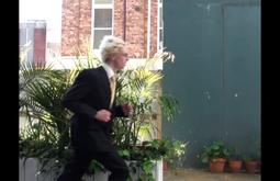 A person wearing a black suit runs across the frame. There are potted plants behind them.