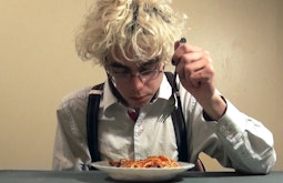 A person wearing a white shirt and suspenders sits at a table eating spaghetti.