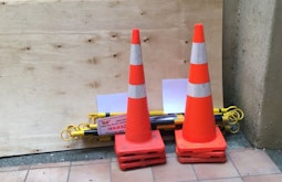 Two road cones and and signs are places by a large sheet of plywood.