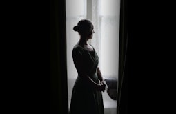 A person stares contemplatively out a window between large dark curtains.