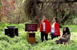 3 people stand in an overgrown field singing Karaoke, there is a TV with lyrics and a speaker beside them.