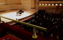 A digitally imposed stainless steel bar runs along the balcony seating in a concert hall. There is a grand piano on the stage in the background.