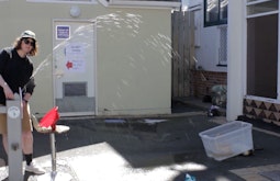 A person stands at a drinking fountain in front of a public toilet spraying the water into a bucket.