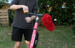 In a backyard someone juices a lemon on the handlebar of an electric scooter. A red cap hangs off the handlebars too.