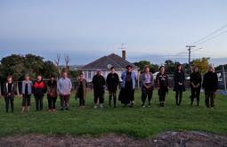 Fourteen people stand in a line on a lawn facing the camera. There is a house behind them.