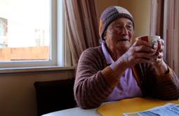 A person wearing a wool beanie and caridgan sits at a table by a window drinking a cup of tea.