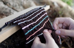 A close up of a weaving being made, the top edge is nailed in place to a plank of wood.
