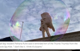 A sculpture of a giant hand with a face stands atop a building. A bubble hovers in front of it. There is a screenshot of text at the bottom of the frame.