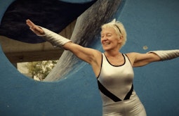 A smiling person wearing a silver leotard and silver arm bands dances with their arms outstretched. There is a large blue sculpture behind them.