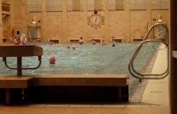 A large indoor pool with many people swimming. The walls are made of orange brick. Bright overhead lights shine down.