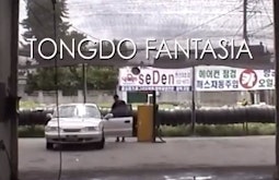 In a car garage a person stands by the open door of a white car. The text "Tongdo Fantasia" is written onscreen.
