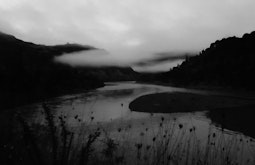 A black and white scene of a river running between dark hills. Low cloud hangs over the river.