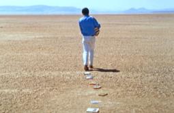 A person walks in a desert carrying an armful of books, some are strewn behind them in a line.