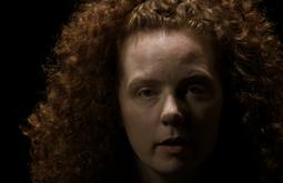 A person looks vacantly into the camera, they're surrounded by black.