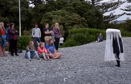 A person wearing large hat like sculpture with long tassels walks along coastal rocks. A group of about 13 people watch on.