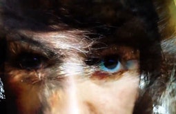 A person close to the camera stares down the lens, blue and yellow hues highlight their face.