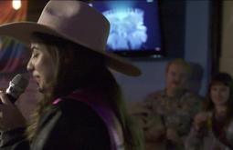 A woman wearing a cowboy hat sings into a microphone. Two audience members sit watching her in the background.